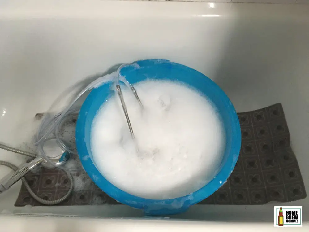 A stainless steel wort chiller soaking in soapy water in a large plastic bowl in the bath